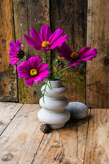A vase with purple flowers sits on a wooden table beside a rock. The flowers are arranged in a way that they are all facing the same direction, creating a sense of harmony and balance