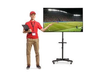 Full length portrait of a shop assistant holding a clipboard and standing next to a tv showing football match