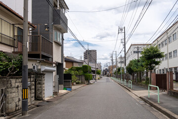 A narrow residential Japanese street lined with low-rise buildings and power lines. A quiet urban road with houses, a school, and a pedestrian sidewalk. A peaceful streetscape in Nagoya, Japan