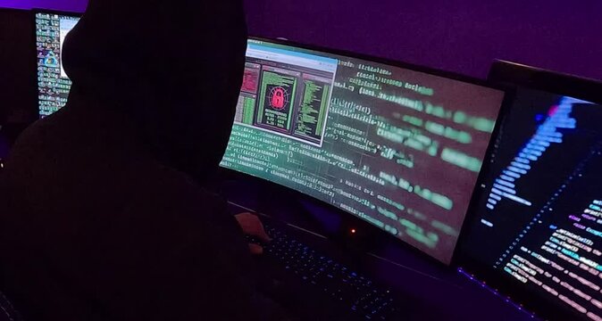 A hooded hacker sits in front of a screen, dragging a window filled with computer system security symbols. The dark atmosphere is highlighted by dim lighting, adding to the sense of secrecy.