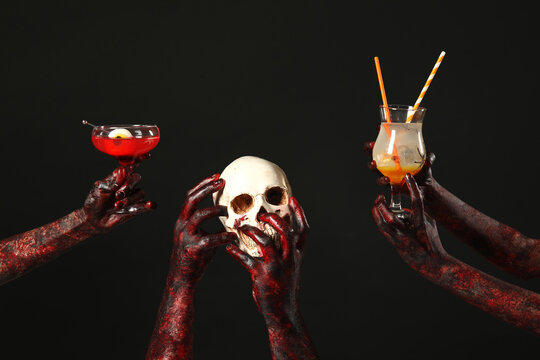 Many hands of zombies with glasses of cocktails and skull on black background. Halloween celebration