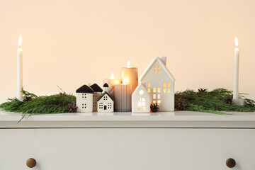 Burning candles with house shaped holders and Christmas branches on commode near light wall