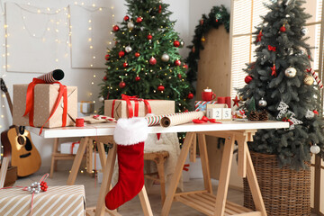 Beautiful gift boxes with packing materials and decorations on table near Christmas trees at home