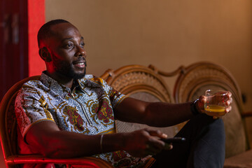 An African man relaxes in a comfortable armchair, holding a remote control and a glass of amber...