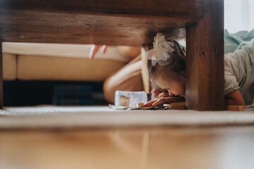 A young child wearing a bow explores under a wooden table, surrounded by toys, in a cozy living room setting. The scene captures innocence, curiosity, and playful moments at home.