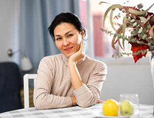 Portrait of positive Asian woman sitting at table in apartment.