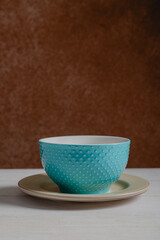 Empty blue and textured blue turquoise ceramic bowl over plate. Wooden white table and brown blurred background.