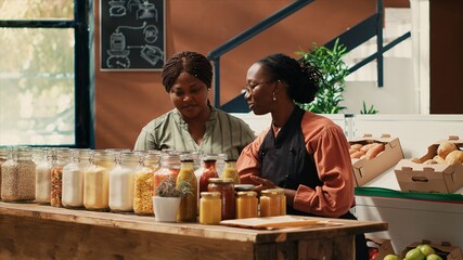 Vendor presenting additives free bulk items for client in store, recommending types of vegan organic pasta in recyclable jars. Merchant with apron showing pantry essentials to buyer, eco friendly.