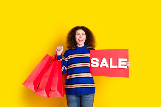 Photo of impressed girl with perming coiffure holding shopping bags sale placard look empty space isolated on yellow color background