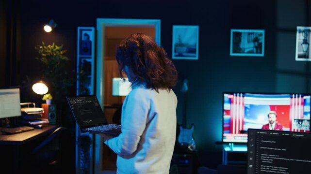 Woman walking in apartment designing and developing software applications using laptop. IT professional standing in living room, implementing and maintaining company systems, camera B