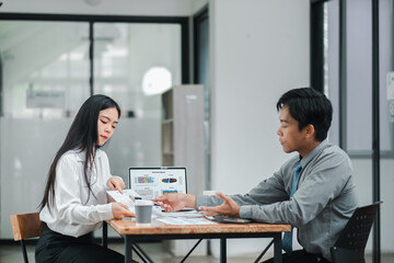Two business professionals in a modern office setting, engaged in a discussion with documents and a laptop displaying data.