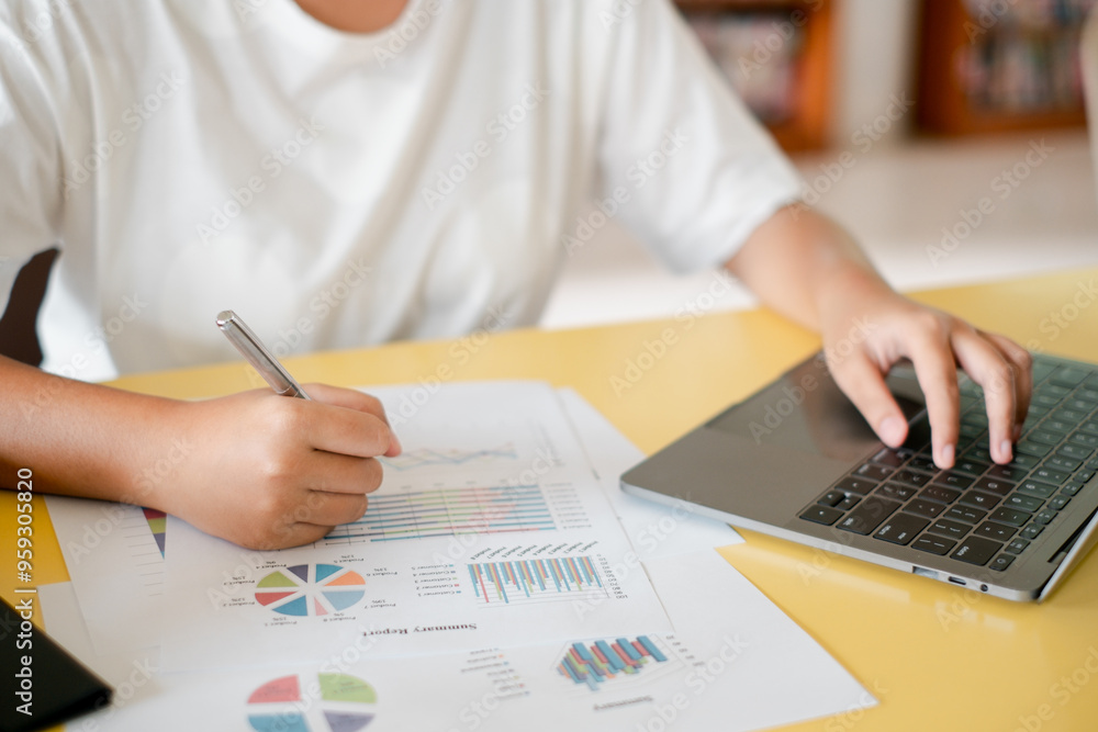 Poster close-up of a person working on financial charts and graphs on paper while using a laptop at a desk.