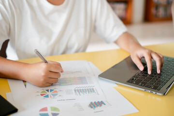 Close-up of a person working on financial charts and graphs on paper while using a laptop at a desk.