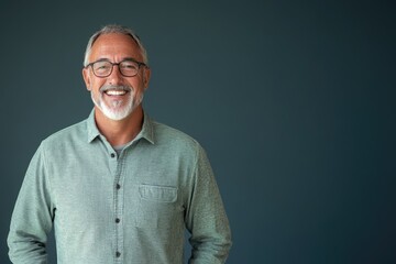 Fototapeta premium Senior male presenter in a casual shirt, smiling, standing against a solid blue-gray background with space for text