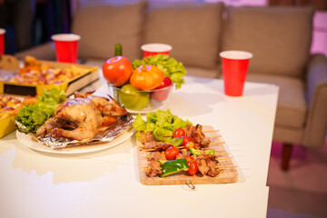 An indoor office party scene with no people around, featuring a desk filled with food pizza, bread, salad, chicken grill, French fries, and beerset against a night light with RGB lighting.