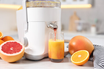 Modern juicer, oranges, grapefruits and glass on white marble table in kitchen