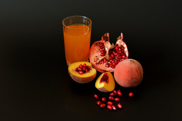 A tall glass of peach-pomegranate juice and pieces of ripe fruit on a black background.