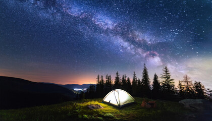 A tent with light in forest on mountain at night with starry milky way galaxy in the sky for outdoor adventure lifestyle, camping product background
