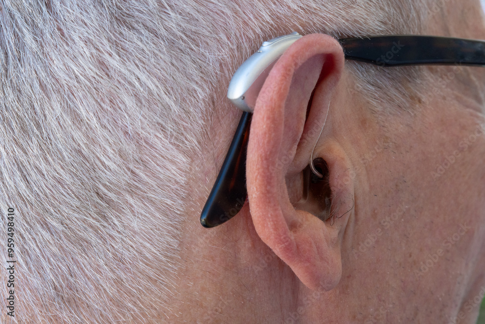 Wall mural close-up of the ear of an elderly grey-haired man wearing glasses and a modern compact hearing aid. 