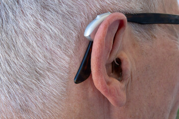 Close-up of the ear of an elderly grey-haired man wearing glasses and a modern compact hearing aid....