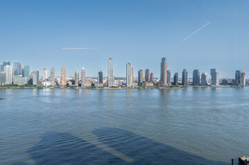 Obraz premium This stunning view of Jersey City, seen across the calm waters of the Hudson River, highlights the towering skyscrapers against a clear blue sky. The modern skyline reflects the vibrant urban landscap
