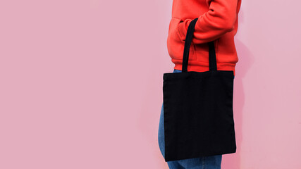 Woman in red hoodie holding tote canvas blank eco bag on street pink minimal wall background. Female consumer hold black textile shopper. template or place for your design, text, logo