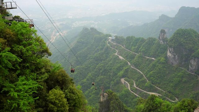 Cable car glide over lush green mountains with winding roads below on a cloudy day. Tianmen Mountain, China