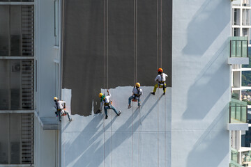 Four workers wearing colourful helmets hanging on ropes at height painting a tall building on a very sunny day.