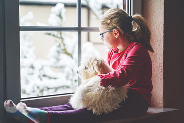 Little girl sitting by window with her pet dog Maltese at home. Happy child and cute puppy looking out on winter snow landscape. Love, friendship, family animal.
