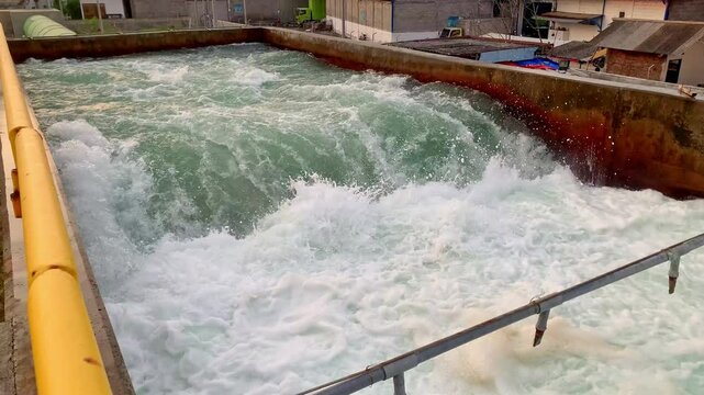 Rapid Water Flow in a Concrete Channel. A view of a water discharge channel, with water rapidly flowing through it. The concrete channel is surrounded by industrial buildings and pipes.