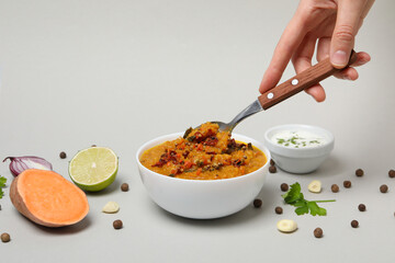 Mashed sweet potatoes with spices in bowl, ingredients and spoon in hand on gray background