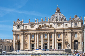 Obraz premium Visitors admire the stunning architecture of St. Peter's Basilica. The grand structure, adorned with sculptures, shines under a bright blue sky, drawing tourists from around the world.