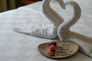 Happy Wedding Anniversary. Two swans made from towel with a cake on a white bed in hotel room.