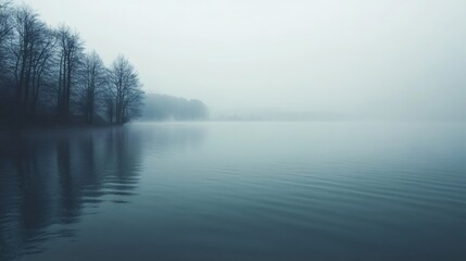 Fototapeta premium Foggy morning on a calm lake with bare trees along the shore.