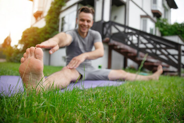 Naklejka premium Man stretches towards his toes while sitting on a mat in a backyard, with one leg extended. The focus is on his foot in the foreground, emphasizing flexibility and an active, healthy lifestyle.