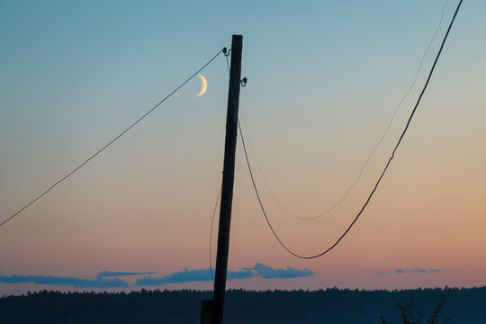 Old leaning wooden pole with sagging electric wires. There is evening sky with pink light and moon. Background.Landscape.
