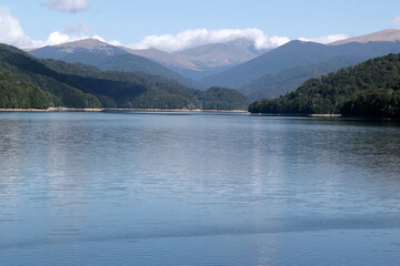 Fototapeta premium Landscape photo with a view of the mountains and the smooth mirror surface of Lake Vidraru against a blue sky with clouds in Romania 