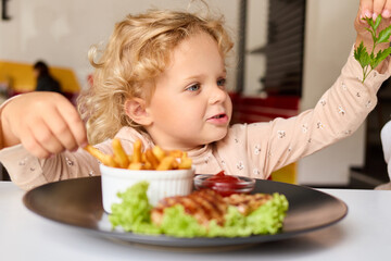 Unhealthy lunch for cute wavy haired blonde toddler girl who is sitting at table eating potato free with grilled meat and sauce looks curious to eat in cafe