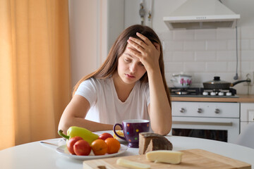 Distraught depressed Caucasian woman sitting at table in kitchen with leaning head on her hand...