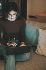A young woman working late at night on her laptop, sitting on a cozy sofa in a dimly lit room....