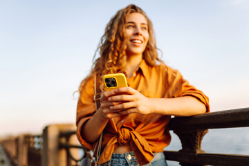 Fototapeta premium Young woman using smartphone by the waterfront during sunset, wearing a casual outfit while enjoying the warm evening light. Lifestyle, travel, nature, active life.