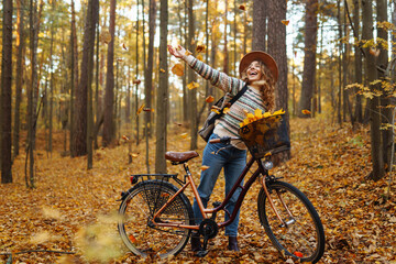 Fototapeta premium A woman biking through a golden autumn forest while enjoying a basket full of vibrant colorful leaves in the late afternoon sunlight. Active lifestyle.