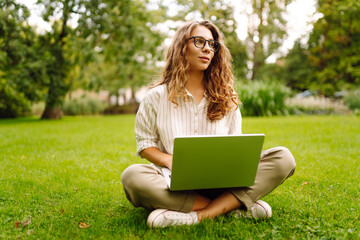 A young woman with curly hair sits comfortably on the grass, using her laptop outdoors. Business, freelancer, online education.