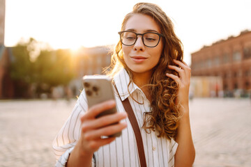 A young woman with curly hair and glasses thoughtfully using her smartphone in an urban plaza...