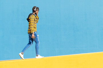 young black man with headphones walking along a yellow wall and a blue wall in the background with...