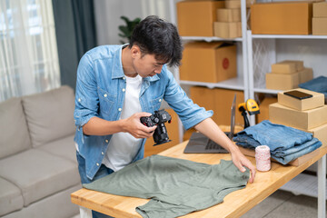 Young Asian man, distributor, online shop owner, small business owner, standing in warehouse, using camera to take pictures of products for sale.