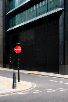A street corner featuring a red no entry sign, against a backdrop of dark modern buildings, highlighting the urban landscape and interplay between infrastructure and architecture.