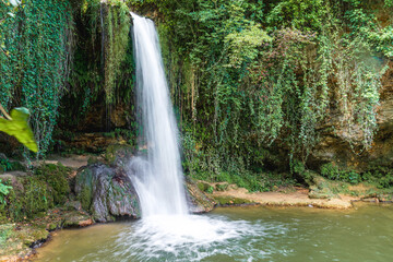 A serene waterfall cascades over rocks into a tranquil pool surrounded by lush greenery in a peaceful outdoor setting