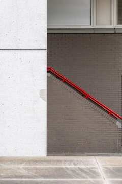 A vibrant red handrail ascends along a brown brick wall, bordered by a large gray structure to the left, creating a contrast in an architectural setting.