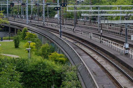 Curved railway tracks lead through an industrial environment, surrounded by lush green vegetation, intricate overhead wires, and various signaling equipment.